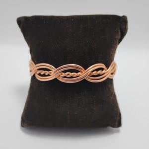 May include: A copper-colored bracelet with an intricate woven design. The bracelet is displayed on a dark brown velvet cushion against a white background. The woven pattern creates a textured, eye-catching accessory.