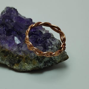 May include: A copper-colored ring with a braided design. The ring is resting on a purple and gray crystal formation. The ring's design features multiple strands of copper wire woven together to create a textured, circular band. The crystal has a rough, natural appearance.