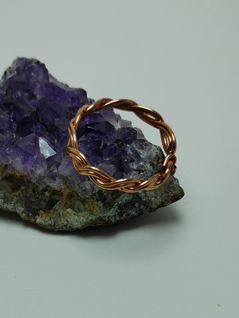 May include: A copper-colored ring with a braided design. The ring is resting on a purple and gray crystal formation. The ring's design features multiple strands of copper wire woven together to create a textured, circular band. The crystal has a rough, natural appearance.