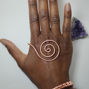 K&ouml;nnte beinhalten: Kupferspiralring und zwei Kupferarmb&auml;nder. Der Spiralring ist auf einer Hand platziert. Ein Armband hat ein gewebtes Design, und das andere ist ein einfaches Band. Ein violetter Amethystkristall befindet sich im Hintergrund.