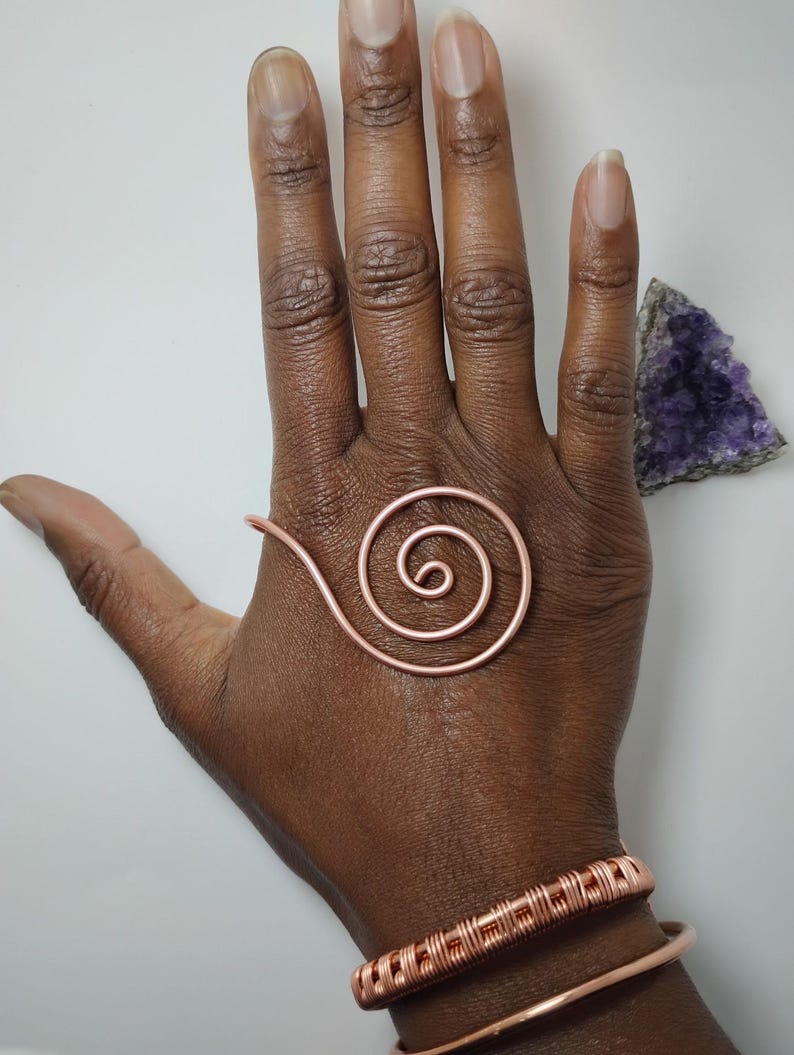 K&ouml;nnte beinhalten: Kupferspiralring und zwei Kupferarmb&auml;nder. Der Spiralring ist auf einer Hand platziert. Ein Armband hat ein gewebtes Design, und das andere ist ein einfaches Band. Ein violetter Amethystkristall befindet sich im Hintergrund.