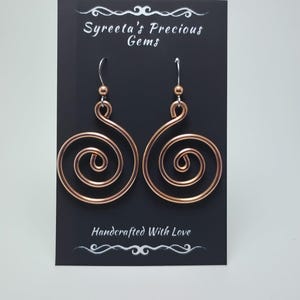 May include: Copper spiral earrings on a black card. The earrings are handcrafted with a spiral design. The card reads "Syreeta's Precious Gems" and "Handcrafted With Love". The earrings have a small copper bead at the top.