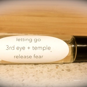 Puede incluir: Un frasco de vidrio transparente con rodillo y tapa negra. La etiqueta dice "letting go 3rd eye + temple. release fear."