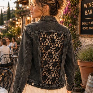 May include: A charcoal gray denim jacket featuring a large, intricate black floral lace panel on the back. The jacket has a classic collar, long sleeves with buttoned cuffs, and a button-front closure. It is worn over a long, cream-colored skirt.