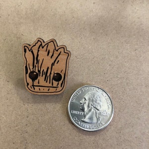 以下が含まれることがあります： ガーディアンズ・オブ・ギャラクシー映画シリーズのキャラクター、グルートの木製切り抜き。切り抜きは茶色で黒いディテールがあり、アメリカの25セント硬貨と同じくらいの大きさです。