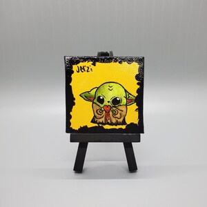May include: A small canvas painting on an easel featuring a cartoon illustration of a green character with large eyes holding a red heart. The background is yellow and the canvas is black with the word "Jerz" written in white.