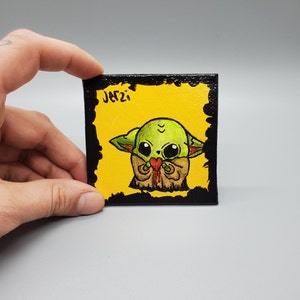 May include: A hand-painted canvas featuring a green cartoon character holding a red heart. The character has large eyes and is wearing a brown outfit. The canvas has a black border and the text "JATZI" in black.