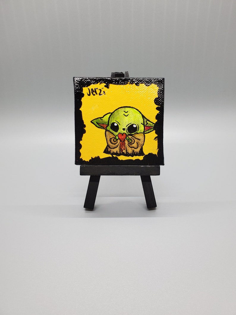 May include: A small canvas painting on an easel featuring a cartoon illustration of a green character with large eyes holding a red heart. The background is yellow and the canvas is black with the word "Jerz" written in white.