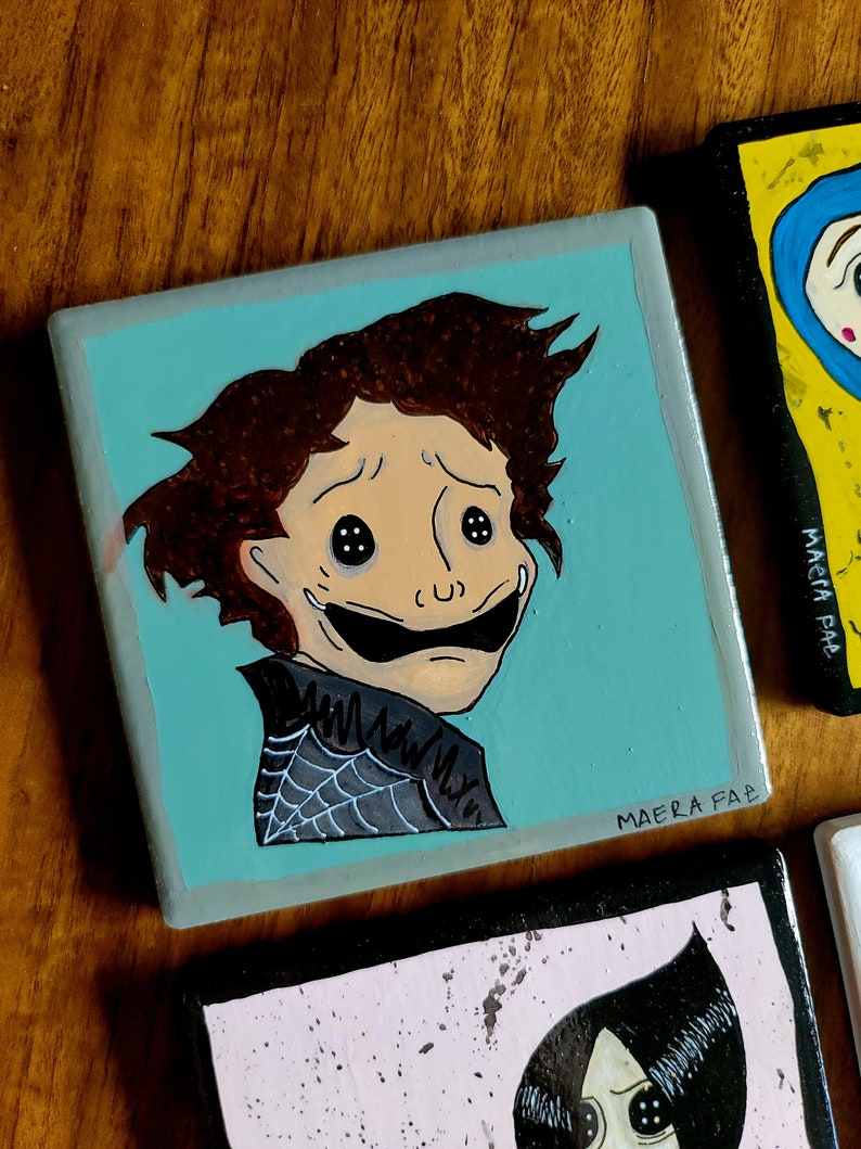 Peut inclure: Un carreau de c&eacute;ramique peint &agrave; la main repr&eacute;sentant un personnage de dessin anim&eacute; avec des cheveux bruns, de grands yeux et un large sourire. Le personnage porte une chemise noire avec un motif de toile d'araign&eacute;e. Le carreau a un fond bleu clair et la signature de l'artiste "MAERA FAP" dans le coin inf&eacute;rieur droit.