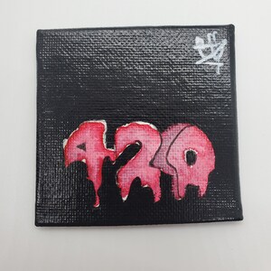 May include: A small black canvas painting with a pink and white dripping paint design that says "420" on it. The painting is signed with a white symbol in the top right corner.