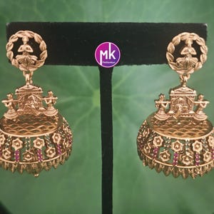 May include: A pair of gold-colored, ornate earrings. Each earring features a circular top with a figure inside a wreath, connected to a detailed, bell-shaped bottom. The bottom has floral and geometric cutouts with green and red accents.
