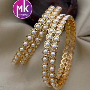 May include: Three gold bangles with white pearl accents. The bangles are stacked on top of each other.
