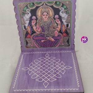 May include: A purple wooden folding stand featuring a printed illustration of a deity seated between two elephants on the upright panel and a white geometric kolam pattern on the base. The edges of the stand have a scalloped design.