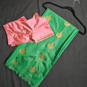 May include: A green silk saree with gold leaf designs and a matching pink blouse. The saree is draped over a hanger.