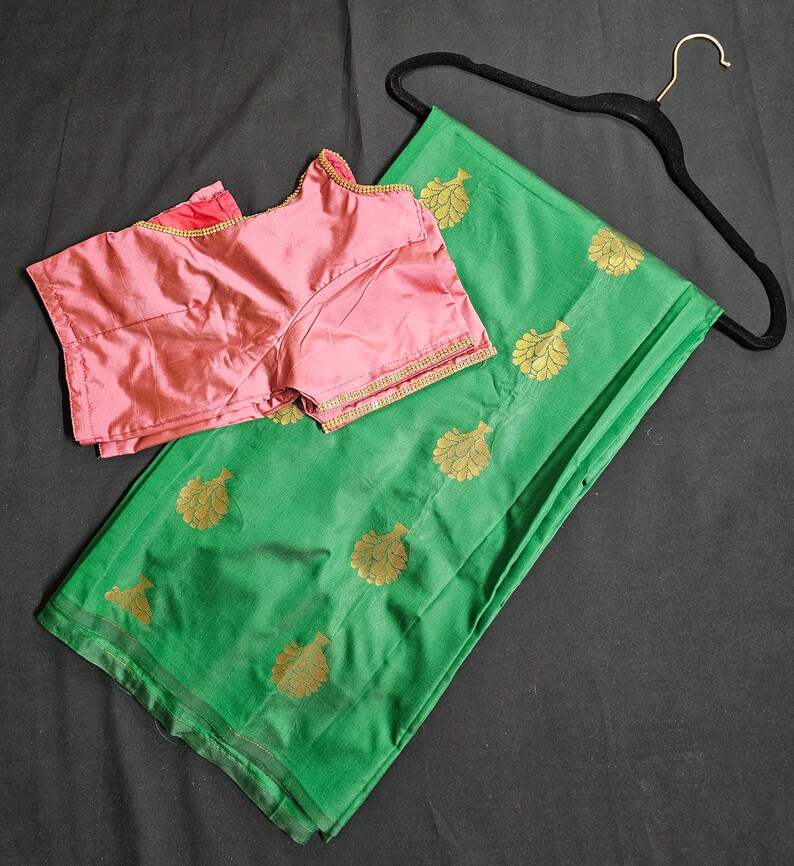 May include: A green silk saree with gold leaf designs and a matching pink blouse. The saree is draped over a hanger.
