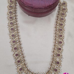 May include: A long gold-toned necklace and matching pair of drop earrings featuring teardrop-shaped maroon stones, clear crystals, and small white pearl accents. The jewelry set is arranged with the necklace forming a U-shape and the earrings above.