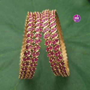 May include: A set of two gold-toned bangles featuring a repeating S-shaped metal pattern embedded with small, round, vibrant pink stones. The bangles have a polished finish and a circular design. A small circular logo with the letters mk is visible.