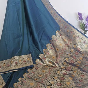Puede incluir: Un sari de seda azul turquesa con un borde dorado y negro. El sari tiene un dise&ntilde;o tradicional indio con intrincados patrones florales y motivos paisley.