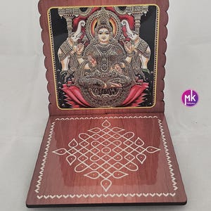 May include: A wooden folding stand with a scalloped edge featuring a colorful illustration of a deity seated on a lotus flower flanked by two elephants. The base displays a white geometric kolam pattern on a brown wood-grain surface.