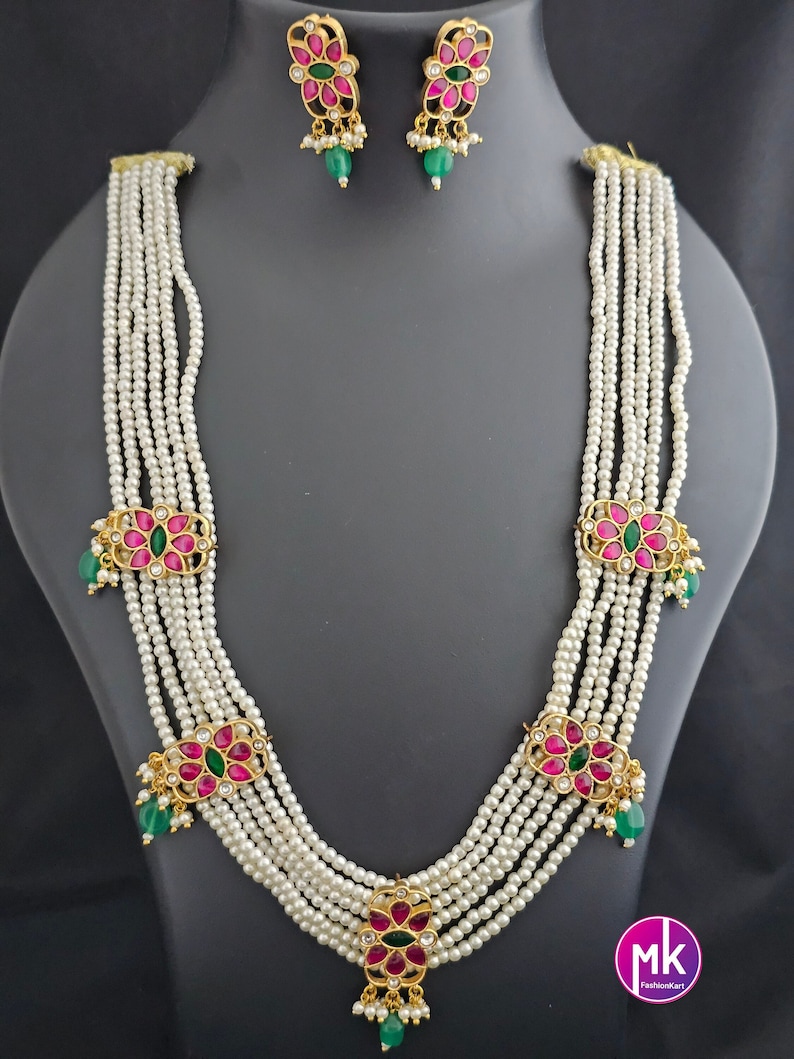 May include: A six-strand pearl necklace with pink and green floral accents. The necklace has five decorative pendants with pink and green stones. The necklace is paired with matching earrings.