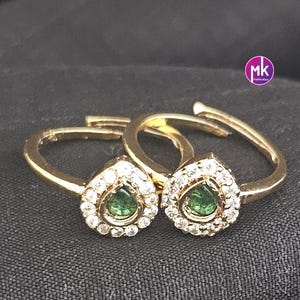 May include: Two gold-toned toe rings. Each ring features a teardrop-shaped setting with a green gemstone center, surrounded by small, clear stones. The rings are displayed on a dark, textured surface.