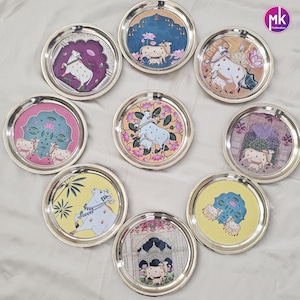 May include: A collection of eight circular stainless steel plates, each featuring a unique, colorful illustration of cows in various settings, including floral patterns and architectural arches, with a small circular logo reading mk in the corner.