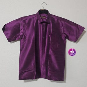 May include: A purple short-sleeved button-down shirt with a pocket on the left side. The shirt has a subtle sheen and a textured fabric.