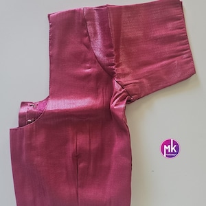 May include: A magenta blouse with short sleeves. The garment has a shiny, satin-like texture and a round neckline. The blouse is laid flat on a white surface. A small, circular logo is visible on the right side.