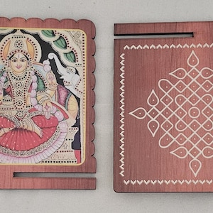 May include: Two square wooden plaques with a dark brown finish. The left plaque features a colorful illustration of a seated deity flanked by two elephants. The right plaque displays a white geometric kolam pattern and a circular mk logo.