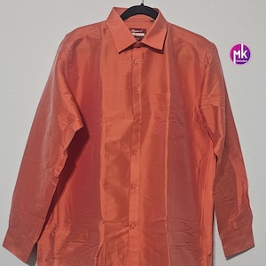 May include: A long-sleeved, coral-colored button-down shirt. The shirt has a classic collar, a chest pocket, and a button-up front. The fabric has a subtle sheen, and the shirt is hanging on a black hanger.