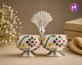 May include: A silver-toned double bowl set featuring a central peacock figure with an open fan tail. Each bowl has a rounded base and is decorated with embossed floral patterns in red, green, and yellow enamel accents on a textured surface.