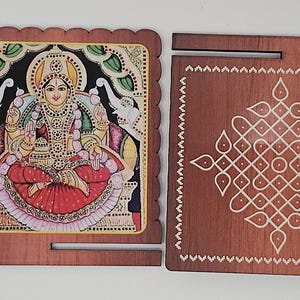 May include: Two square wooden plaques with scalloped edges. One features a colorful illustration of a deity flanked by elephants, measuring 4 inches wide. The other is a dark wood plaque with a white geometric kolam pattern and a slot.