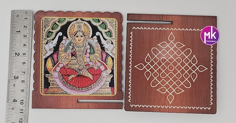 May include: Two square wooden plaques with scalloped edges. One features a colorful illustration of a deity flanked by elephants, measuring 4 inches wide. The other is a dark wood plaque with a white geometric kolam pattern and a slot.