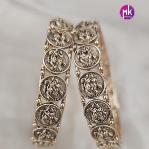May include: Two gold-colored bangles with detailed, circular medallions featuring figures in relief. The bangles are displayed against a soft, white background. The medallions are connected by small, round beads.