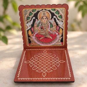 Peut inclure: Un support en bois à bord festonné présentant une illustration colorée d'une divinité assise sur un lotus, flanquée de deux éléphants blancs. La base est une surface brune polie et plate ornée d'un motif géométrique kolam blanc.