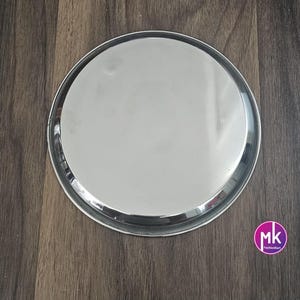 May include: A round, silver serving tray with a smooth, reflective surface. The tray is made of stainless steel and has a slightly raised rim.