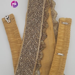 May include: Mustard yellow fabric with intricate gold beaded embellishments. The design includes a central beaded strip with a scalloped edge, and two matching fabric straps with metal clasps. The logo "MK FashionKart" is visible.