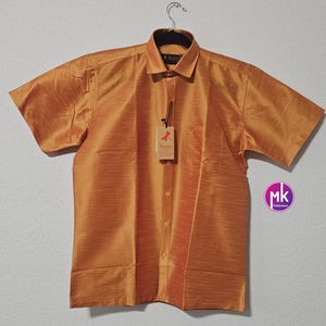 May include: A short-sleeved, gold-colored button-down shirt with a pocket on the left chest. The shirt has a subtle sheen and a textured fabric. The brand name "Azotice" is visible on the tag.