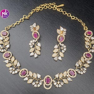 May include: A gold-toned necklace and earring set. The necklace and earrings feature oval, ruby-colored stones surrounded by clear, sparkling accents. The design incorporates leaf and floral motifs.