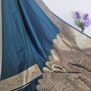 Puede incluir: Un sari azul turquesa con un borde dorado y negro. El borde presenta intrincados patrones florales y geom&eacute;tricos. El sari est&aacute; hecho de una tela suave y lujosa.