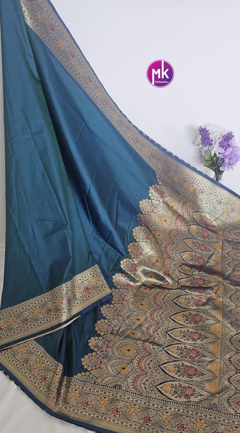 Puede incluir: Un sari azul turquesa con un borde dorado y negro. El borde presenta intrincados patrones florales y geom&eacute;tricos. El sari est&aacute; hecho de una tela suave y lujosa.