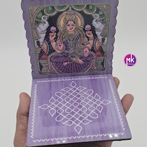 May include: A purple wooden folding stand featuring a printed illustration of the Hindu goddess Lakshmi seated between two elephants on the top panel and a white geometric kolam pattern on the bottom panel. The edges have a scalloped design.