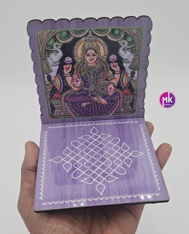 May include: A purple wooden folding stand featuring a printed illustration of the Hindu goddess Lakshmi seated between two elephants on the top panel and a white geometric kolam pattern on the bottom panel. The edges have a scalloped design.