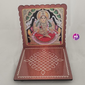 May include: A wooden folding stand with a scalloped edge, featuring a colorful illustration of a deity seated on a throne with two elephants on the sides. The base displays a white geometric kolam pattern on a polished brown wood surface.
