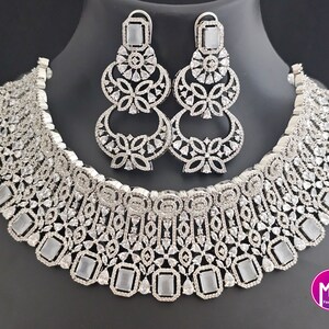 May include: A silver necklace and earrings set with a white stone design. The necklace has a wide, layered design with rectangular stones and smaller stones. The earrings are dangling and have a similar design to the necklace.