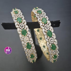 May include: Two circular bangles featuring a metallic finish with embedded oval-shaped green stones and small clear stones arranged in a floral-inspired pattern. A circular logo with the letters mk is visible in the lower left corner of the image.