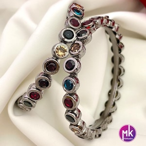 May include: Two silver bangles with a row of colorful round stones. The stones are red, blue, green, yellow, and black.