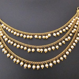 May include: Gold chain with three strands of white pearls hanging from it. The chain has a hook clasp.
