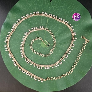 May include: A gold-toned chain belt featuring a series of small white beads dangling along the length of the chain. The belt includes a circular link extension chain at one end and a hook clasp for closure, with a small circular logo tag present.