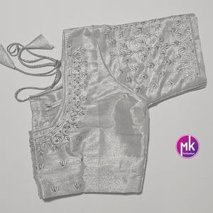 May include: A silver-colored blouse with intricate embroidery along the neckline, sleeves, and bodice. The blouse features a tie closure and decorative tassels. The fabric has a subtle sheen, and the design includes floral patterns. The blouse is a traditional garment.